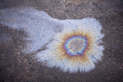 Close-up of an iridescent oil or gasoline spill on a wet asphalt, viewed from above. Bold multicolored spots on the asphalt.