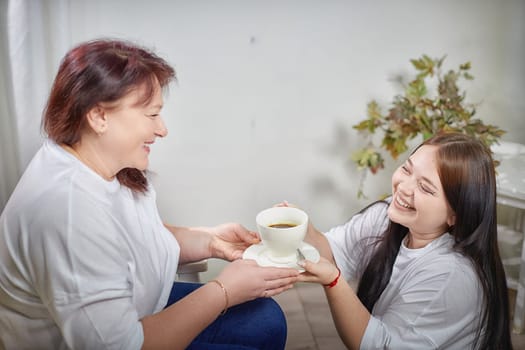 Happy Overweight family with mother and daughter drinking tea or coffee in room. Middle aged woman and teenager girl having fun and joy