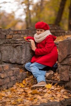 Sad caucasian girl in a red coat and beret sits on a brick wall on a walk in autumn
