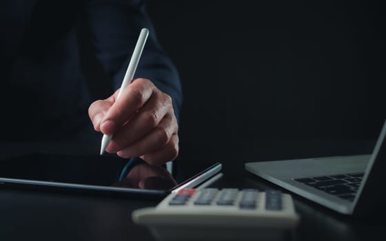 Businessman in black suit working on laptop computer and tablet and calculator, Hand Hands touch on tablet at office with dark background, Online working, Close up, Copy space.