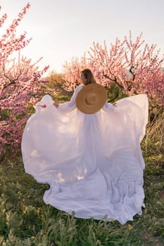 Woman blooming peach orchard. Against the backdrop of a picturesque peach orchard, a woman in a long white dress and hat enjoys a peaceful walk in the park, surrounded by the beauty of nature