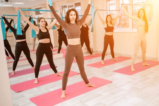 A group of six athletic women doing pilates or yoga on pink mats in front of a window in a beige loft studio interior. Teamwork, good mood and healthy lifestyle concept