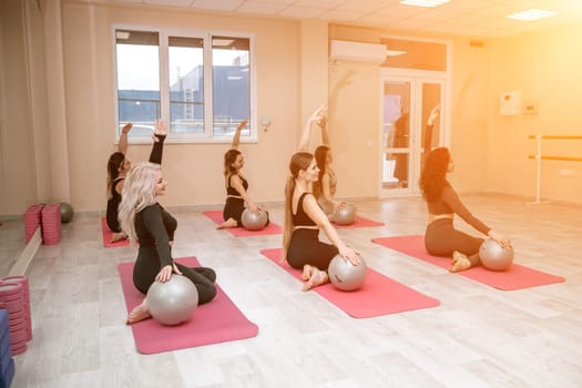 A group of six athletic women doing pilates or yoga on pink mats in front of a window in a beige loft studio interior. Teamwork, good mood and healthy lifestyle concept