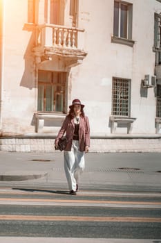 Woman city road crossing. Stylish woman in a hat crosses the road at a pedestrian crossing in the city. Dressed in white trousers and a jacket with a bag in her hands