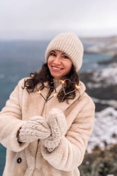 Outdoor winter portrait of happy smiling woman, light faux fur coat holding heart sparkler, posing against sea and snow background.