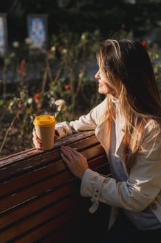 Woman drinks from cup on wooden bench. She is wearing a white shirt enjoying her beverage. The bench is located in a park setting, with trees in the background