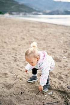 Little girl squats on the beach and draws in the sand with a stick. High quality photo