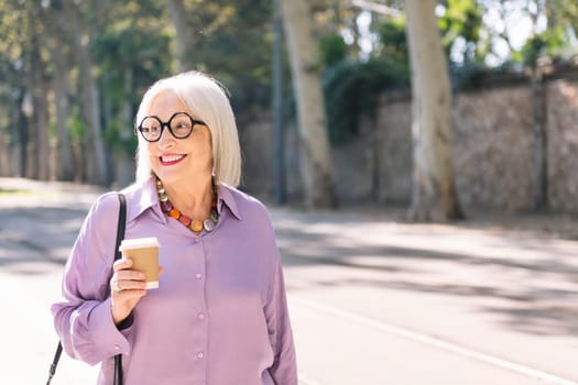 portrait of a beautiful senior woman walking on the street holding a takeaway coffee in her hand, concept of elderly people leisure and active lifestyle, copy space for text