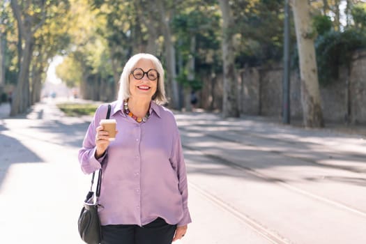 senior woman walking on the street holding a takeaway coffee in her hand, concept of elderly people leisure and active lifestyle, copy space for text