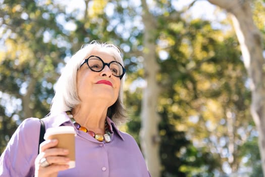 portrait from below of a beautiful senior woman walking outdoors holding a takeaway coffee in her hand, concept of elderly people leisure and active lifestyle, copy space for text