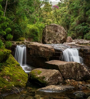 A double waterfall in a rocky river edged with dense jungle, featuring a large, teetering rock and silky water due to long exposure.