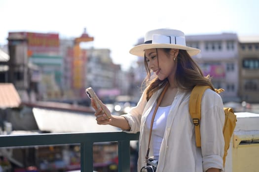 Portrait of smiling female traveller with backpack standing on bridge and using smartphone.
