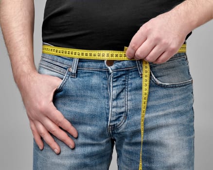 Close-up of yellow tape measure in male hands in casual blue jeans and black t-shirt measuring waistline, cropped shot. Concept of weight control, fitness, tailoring or belt selection