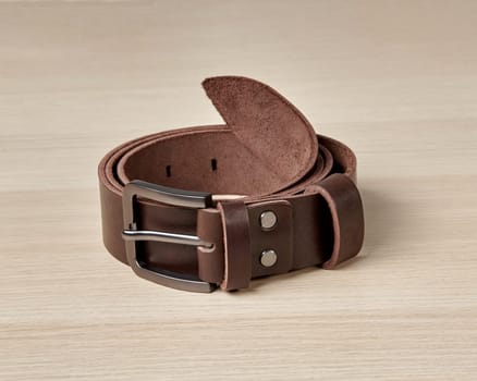 Folded dark brown leather belt with stylish matte buckle and DAD embossing on loop against light wooden background. Stylish personalized handcrafted mens accessory