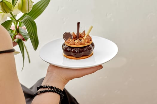 Woman holding white lilies and plate with sophisticated cake with chocolate mousse, shortbread biscuits garnished with caramel peaks and edible gold flakes, cropped image. Exquisite romantic dessert