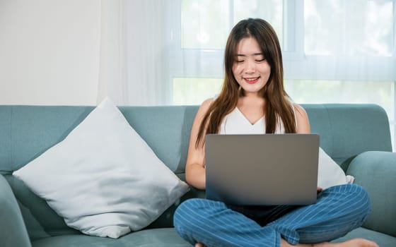 Asian young woman smiling sitting relaxing on sofa using laptop in living room at home, freelance working on computer project, Happy female typing email on notebook computer