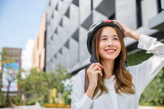 Close up smiling woman wearing helmet exercise outdoors with bicycle on street, Asian young businesswoman putting biking helmet for safety prepared cyclists around building go to work, Bike commuting