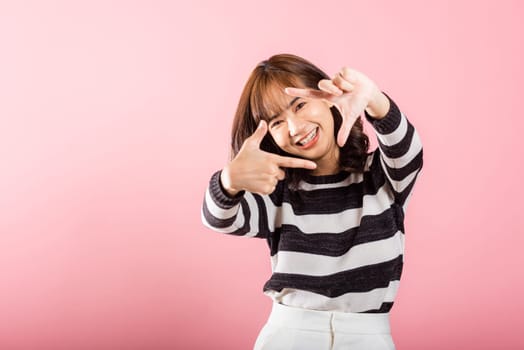 An Asian photographer, with a cheerful smile, uses her finger to create a frame in front of her face for photography. Studio shot isolated on pink background, emphasizing creativity and inspiration.