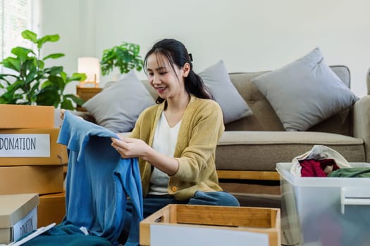 Asian young woman packing clothes at home, putting on stuff into donate box with second hand clothes.