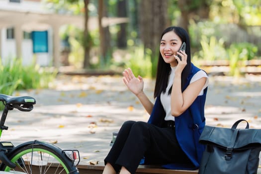 Smiling asian businesswoman hold reusable eco-friendly ecological cup and using mobile while sitting at park.