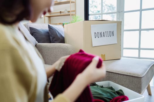 Asian young woman packing clothes at home, putting on stuff into donate box with second hand clothes.