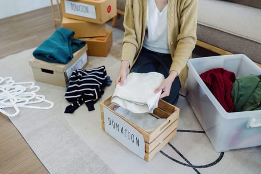 Asian young woman packing clothes at home, putting on stuff into donate box with second hand clothes.