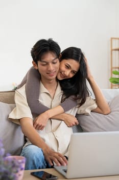 Happy asian couple using laptop sit relax on sofa doing ecommerce shopping online on website at home.