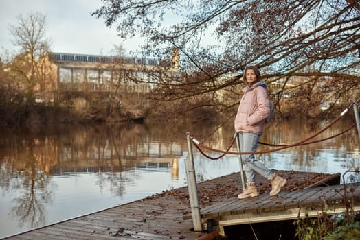 A young woman standing at the shore looking at the river in autumn sunny day. Street view, copy space for text, travel photo. Happy tourist woman on the bank of the river in autumn in warm clothes. Tourists enjoy their vacation, winter season. Romantic look and travel concept. A joyful mood in a Caucasian girl. Winter Wonderland: Enchanting Girl by the Riverside in Autumn.