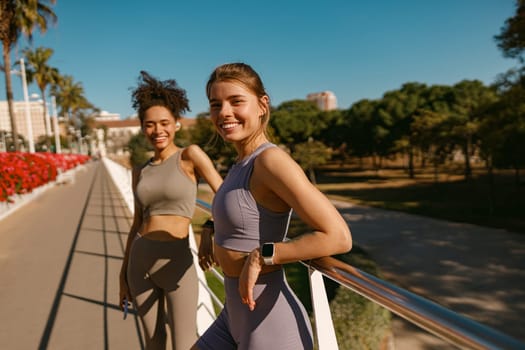 Two young female sportswomen have a rest after morning jogging outdoors and looks camera