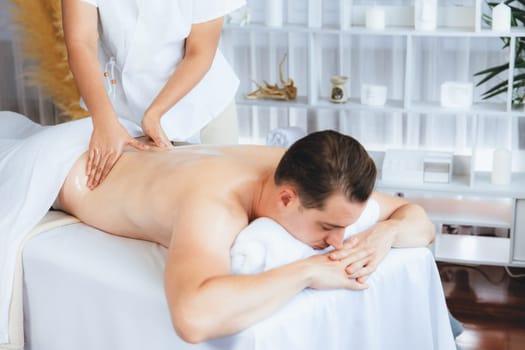 Caucasian man customer enjoying relaxing anti-stress spa massage and pampering with beauty skin recreation leisure in day light ambient salon spa at luxury resort or hotel. Quiescent