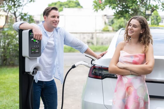 Happy and lovely couple with eco-friendly conscious recharging electric vehicle from EV charging station. EV car technology utilized as alternative transportation for future sustainability. Synchronos