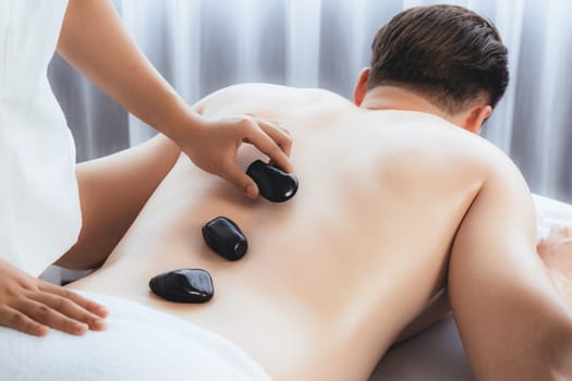 Hot stone massage at spa salon in luxury resort with day light serenity ambient, blissful man customer enjoying spa basalt stone massage glide over body with soothing warmth. Quiescent