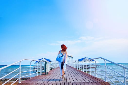 A stunning woman with fiery red hair strolls along the resort pier, adorned in a summer dress and carrying a backpack, with the sea as her picturesque backdrop.