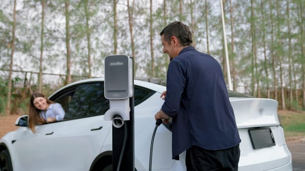 EV car recharging electricity for battery by lovey couple traveling during their road trip travel by eco friendly electric vehicle in national park autumnal forest on holiday. Exalt