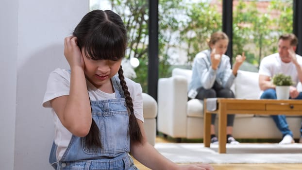 Stressed and unhappy young girl huddle in corner, cover her ears blocking sound of her parent arguing in background. Domestic violence at home and traumatic childhood develop to depression. Synchronos