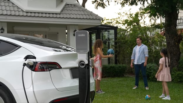 Electric vehicle recharge from home charging station on background of happy and playful family playing together. EV car using alternative and sustainable energy for better future Synchronos