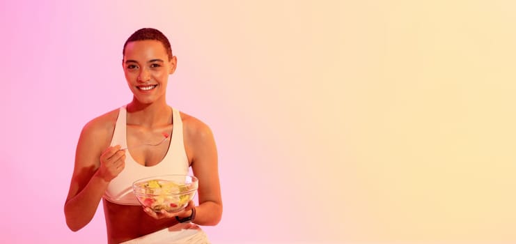 Healthy lifestyle concept with a beaming woman in sports attire, holding a bowl of fresh fruit salad and enjoying a nutritious post-workout snack against a gradient background.