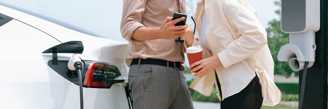 Young couple with coffee cup, recharge electric car's battery from EV charging station in green city park. Sustainable and eco friendly EV car with urban and shopping lifestyle. Panorama Expedient