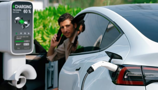 Young man recharge EV electric vehicle at green city mall parking lot while talking on phone. Sustainable urban lifestyle for eco friendly EV car with battery charging station. Panorama Expedient