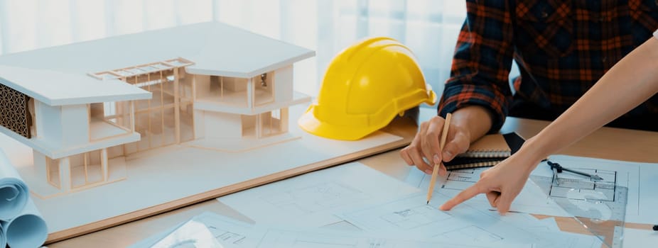 Cropped image of cooperative architect team decide and work together on meeting table with house model, safety helmet and architectural plan scatter around. Closeup. Focus on hand. Burgeoning.