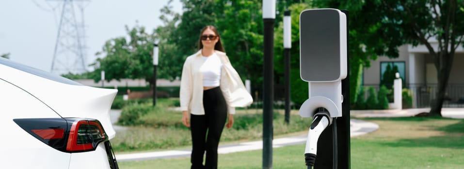 Young woman recharge EV car battery at charging station connected to power grid tower electrical industrial facility as electrical industry for eco friendly vehicle utilization. Panorama Expedient