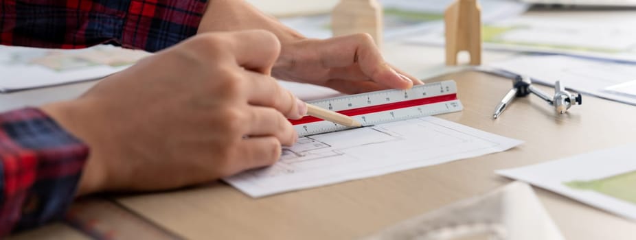 Closeup of architect engineer hand using ruler to mature and draw a blueprint on meeting table with wooden block, pencil and blueprint scatter around at architectural modern office. Delineation.