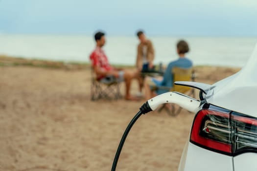 Alternative family vacation trip traveling by the beach with electric car recharging battery from EV charging station with blurred family enjoying the seascape background. Perpetual