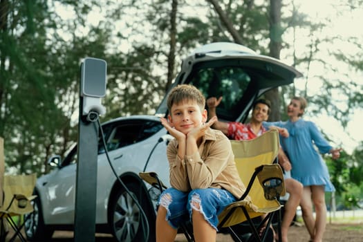 Little boy portrait sitting on camping chair with his family in background. Road trip travel with alternative energy charging station for eco-friendly car concept. Perpetual