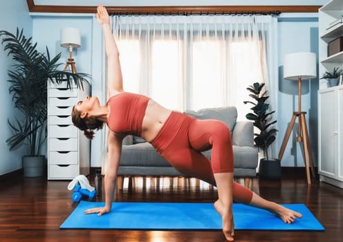 Flexible and dexterity woman in sportswear doing yoga position in meditation posture on exercising mat at home. Healthy gaiety home yoga lifestyle with peaceful mind and serenity.