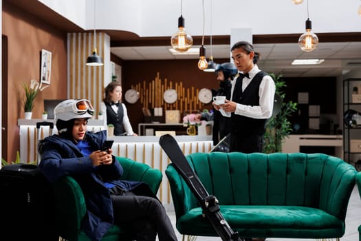 In hotel reception asian female tourist in winter attire uses digital device to explore snow gear and winter activities. Young waiter holding cup of coffee to woman with smartphone in lounge area.