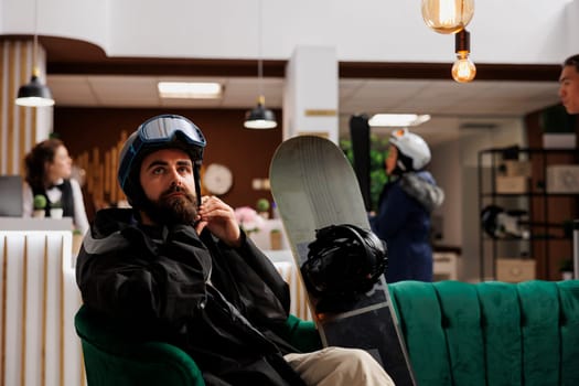 Male tourist in lounge area adjusting ski helmet while waiting for booking process at winter mountain resort. Caucasian man with wintersports gear sitting with snowboarding equipment in hotel lobby.