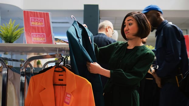 Woman customer looks through clothes hanging on racks, searching for black friday special offers in trendy clothing boutique. Shopper holding items with discount red sale price tags.