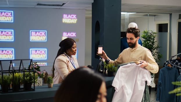 Mobile coupon marketing in retail shop. Client showing smartphone with black friday promo code to cashier, preparing to buy clothes at promotional prices. Shopper using loyalty voucher.