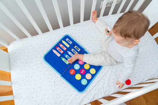 One year old baby playing with montessori busy book sitting in crib. ducational books and quiet books concept. Montessori busy board